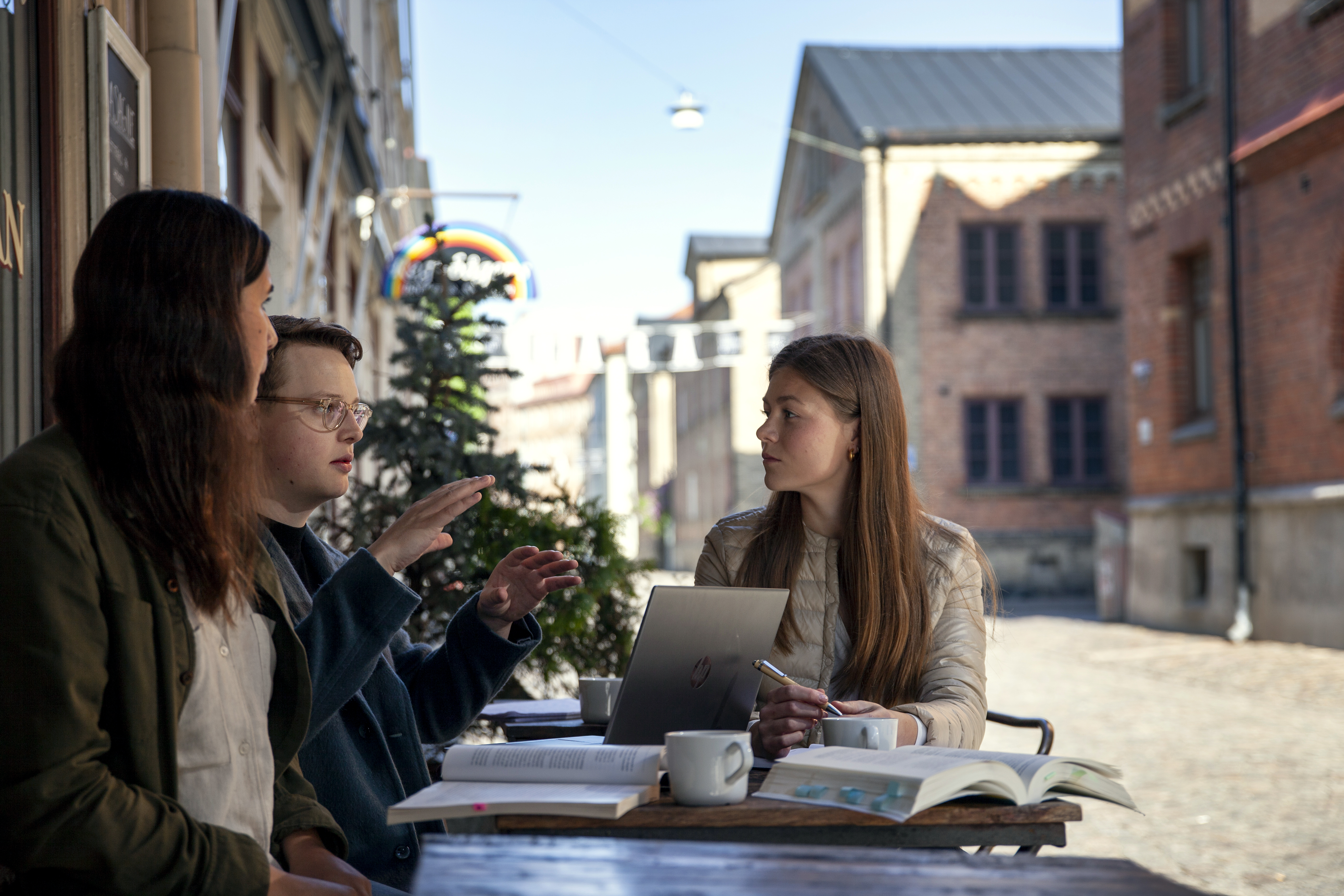 Tre studenter samtalar vid cafebord fyllt med böcker och kaffekoppar.