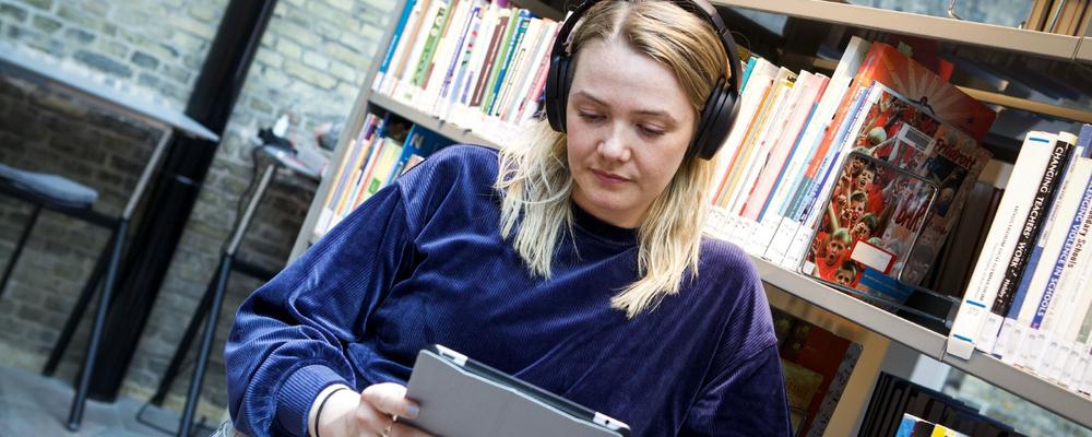Kvinnlig student med hörlurar står i ett bibliotek och håller i en surfplatta.