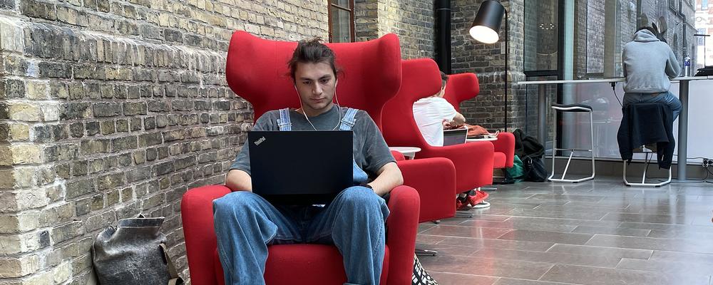Male student sitting in a red armchair with a laptop on his knee, listening to something in headphones.