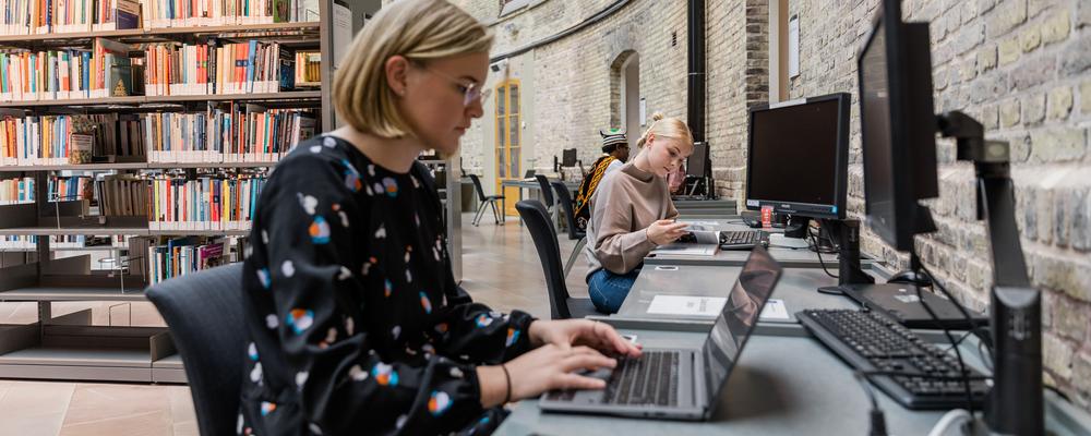 Kvinnlig student sitter vid en dator på biblioteket och tittar in i skärmen.