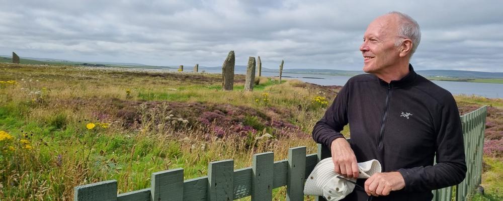 En man med grått hår och svarta funktionskläder står till höger i bild, han ler i profil. Mannen står vid ett grönt trästaket och tittar ut över en äng eller naturreservat med stora lodräta stenar – Ring of Brodgar. Bakom honom syns sjölandskap.